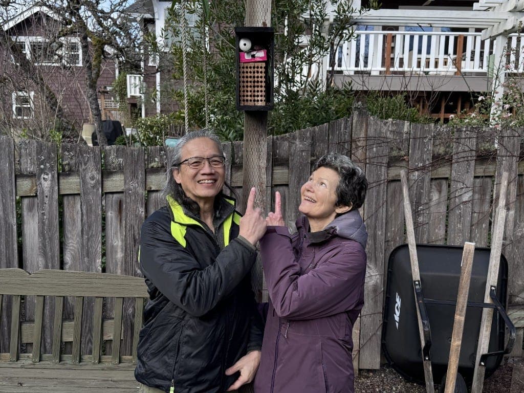 Two hosts pointing at a black bee house hung on a fence post