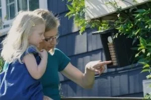A woman holds a child and points at a black mason bee house on blue siding
